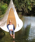 Child sitting in a hanging chair by a body of water with trees in the background