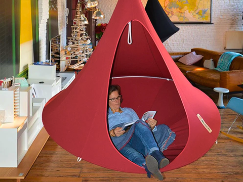 Person reading a book inside a red hanging pod in a modern living room.