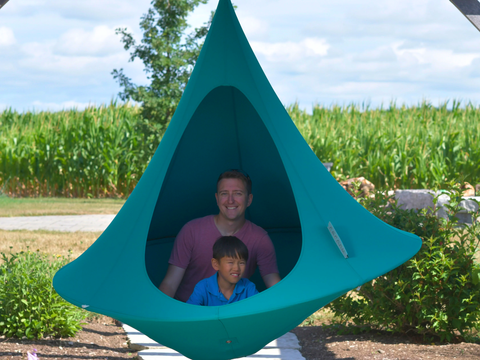Two people inside a teal hanging pod with a cornfield in the background