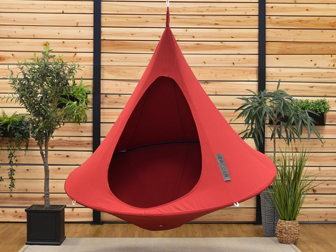 Red hanging pod chair against a wooden wall with plants.