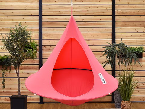 Pink hanging pod chair against a wooden wall with plants.