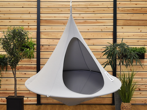 Gray hanging chair with 'Cacoon' branding against a wooden wall with plants.