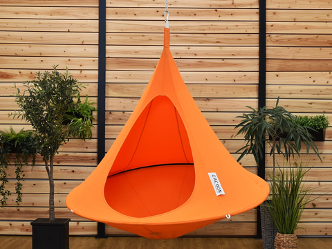 Orange hanging chair with a brand label against a wooden wall with plants.