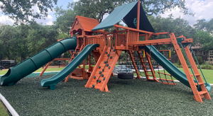 Huge wooden playground on green rubber mulch with tube slide, rock walls, monkey bars, and tire swings.