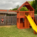 Wooden playset with a yellow slide and swing set in a backyard.