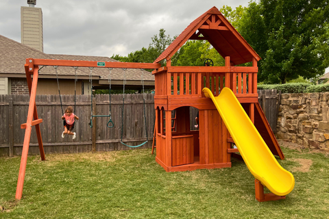 Wooden playset with a yellow slide and swing set in a backyard.
