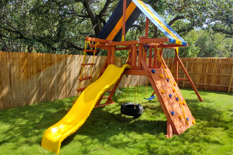 Children's playground set with yellow slide and climbing wall in a backyard.