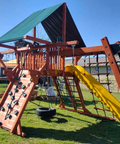 Wooden playground structure with a yellow slide and climbing wall on a grassy area.